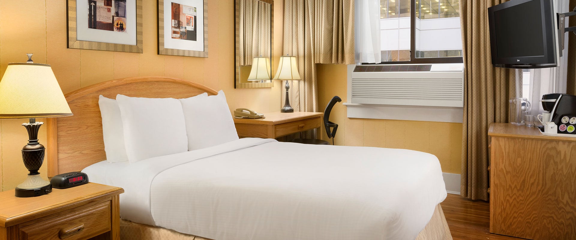 The Standard King room at Days Inn Vancouver Downtown features a neatly made bed in white sheets, natural beige wood colored bedside table and writing desk and air conditioner.