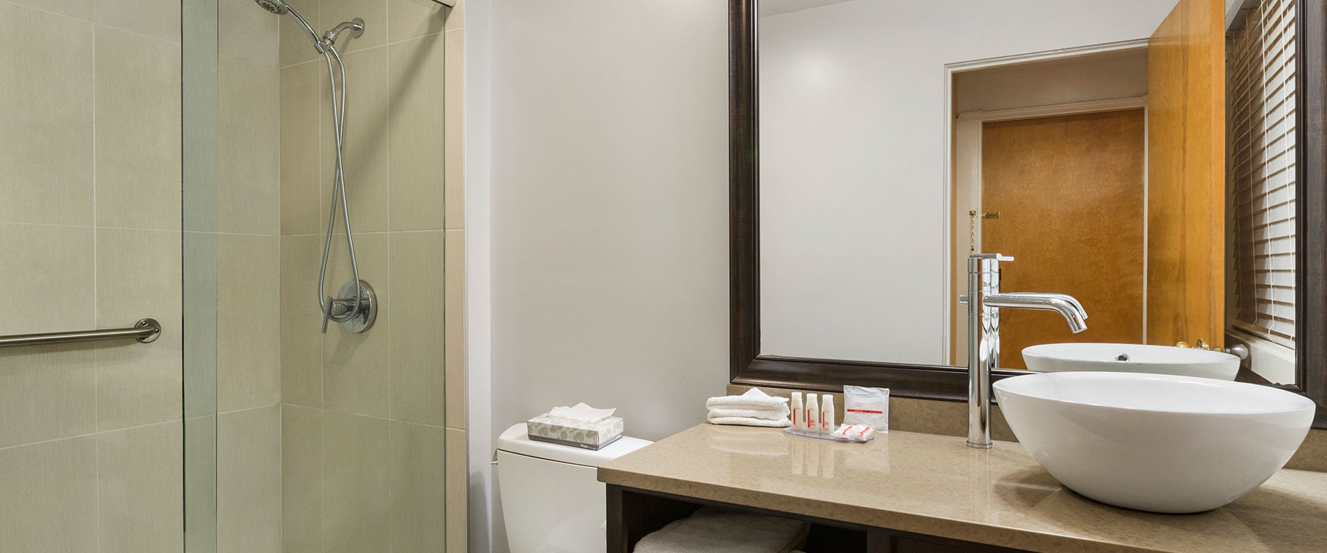A bowl shaped white vessel sink, a beige colored vanity with soaps and towels and beige tiled shower are features in a bathroom suite at Days Inn Vancouver Downtown.