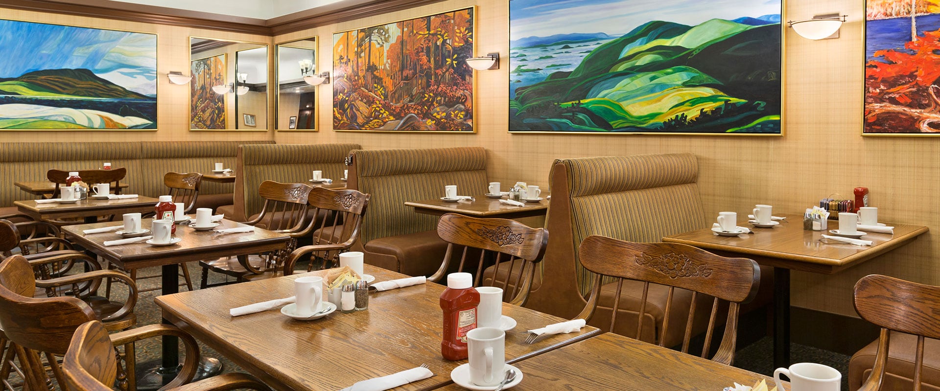 Meals are ready to be served at Chelsea's Restaurant with cups and saucers, eating utensils, bottles of ketchup and condiments set atop brown wood grain dining tables.