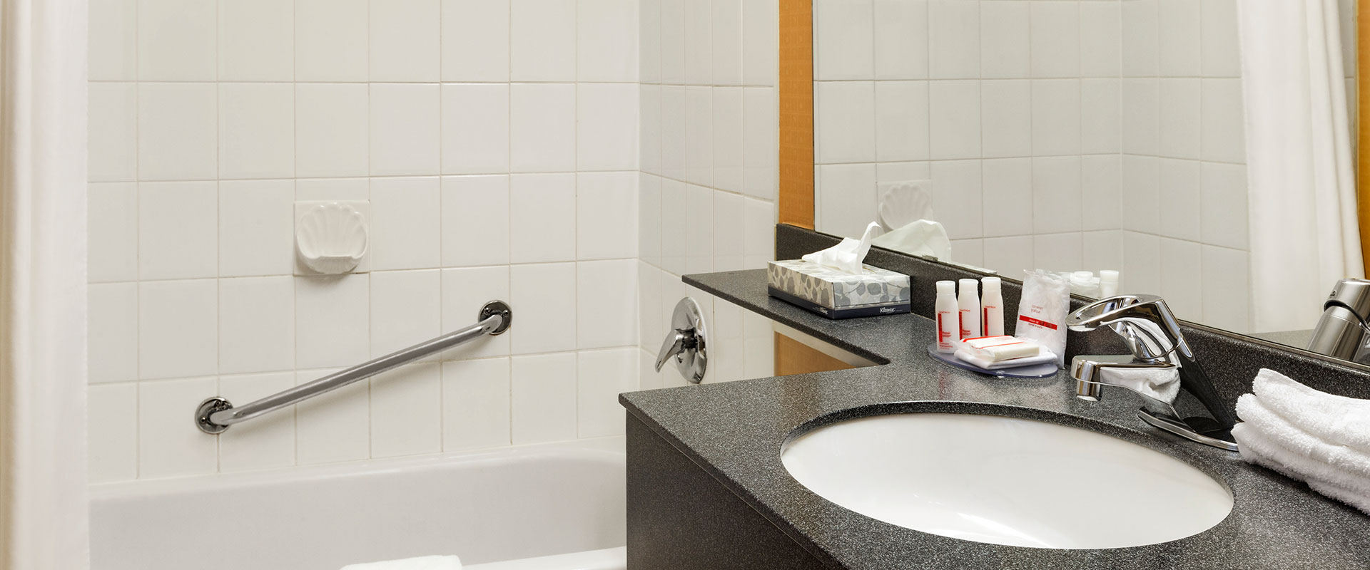 A charcoal colored bathroom sink countertop stocked with soaps, towels and tissue with a large mirror above in a suite at Days Inn Vancouver Downtown.