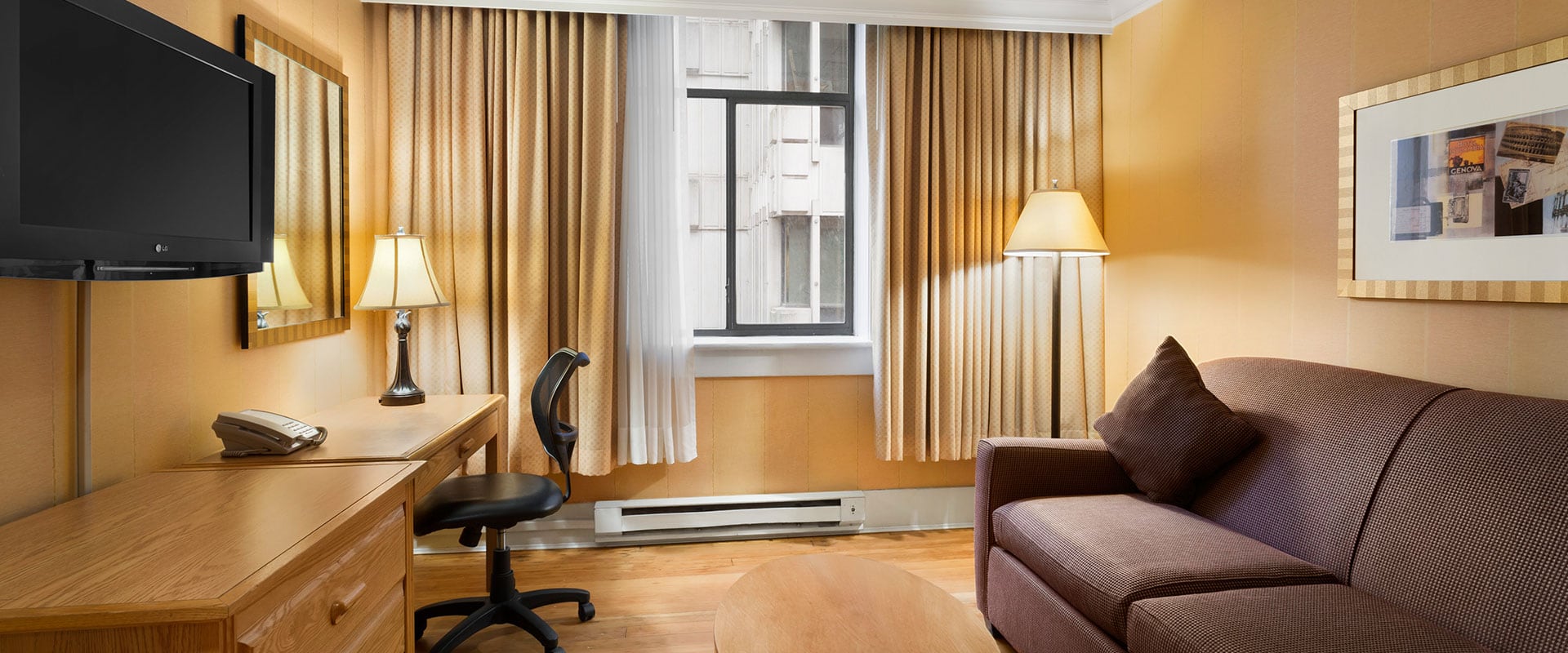 A seating area with a brown loveseat and natural wood colored oval coffee table, writing desk and a mounted flatscreen TV in the Queen Suite at Days Inn Vancouver Downtown.