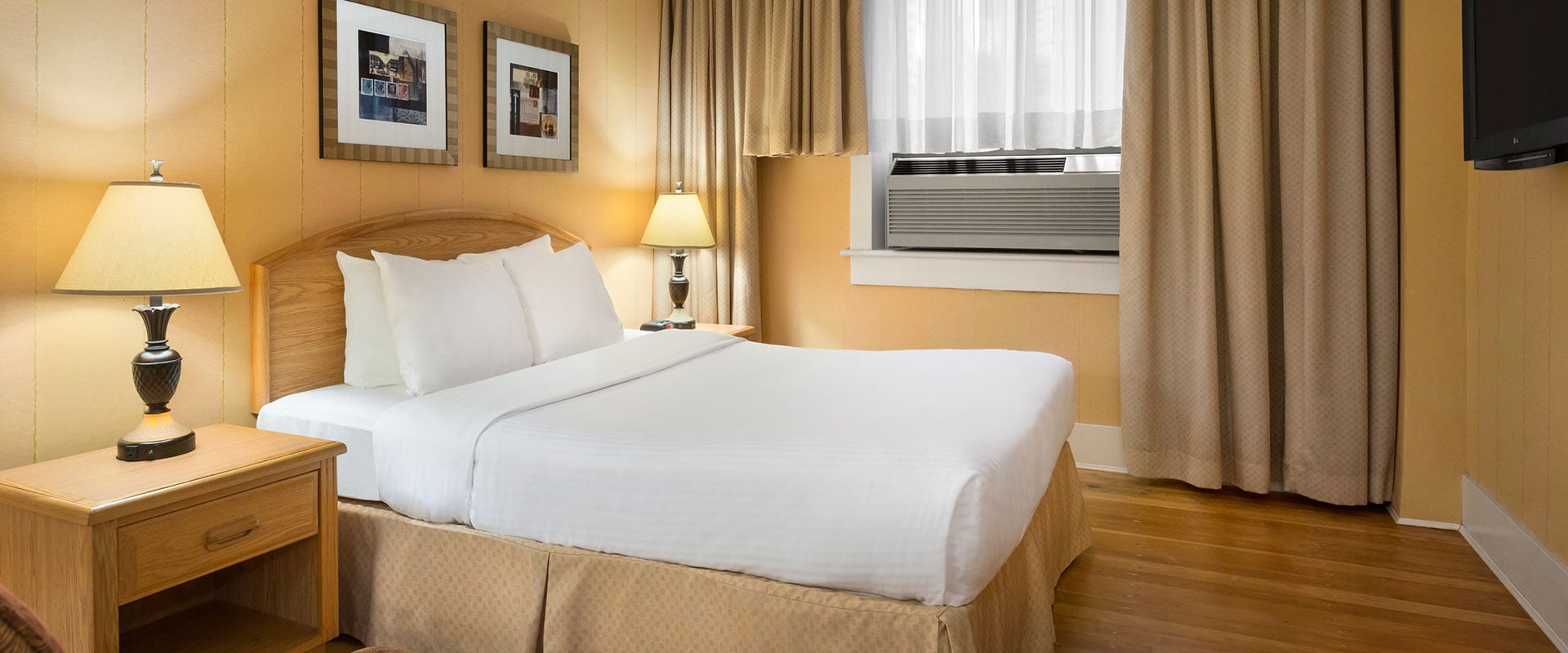 Canary yellow walls, natural wood colored bedside tables, and beige curtains are contrasted against a bed in bright white linen in the Standard Double suite at Days Inn Vancouver.