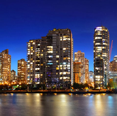 The lights from tall skyscapers of Vancouver Dowtown reflect in the dark waters of a shoreline and against the dark blue night sky.