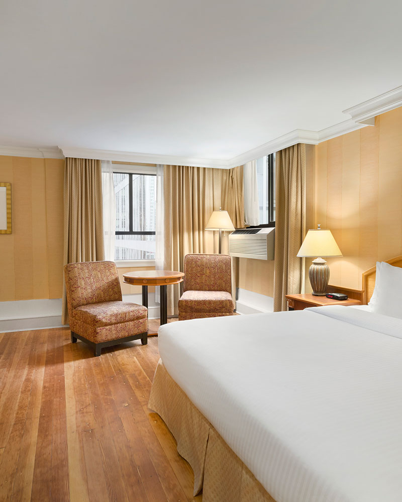 The Standard Queen suite at Days Inn Vancouver Downtown with a large bed in white linen, hard wood flooring, two patterned chairs and a round table placed by the window.