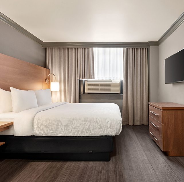Two framed pictures hang above a bed in the Standard Queen suite at Days Inn Vancouver furnished with a bedside table and a round wood-topped table and two chairs placed by a window.