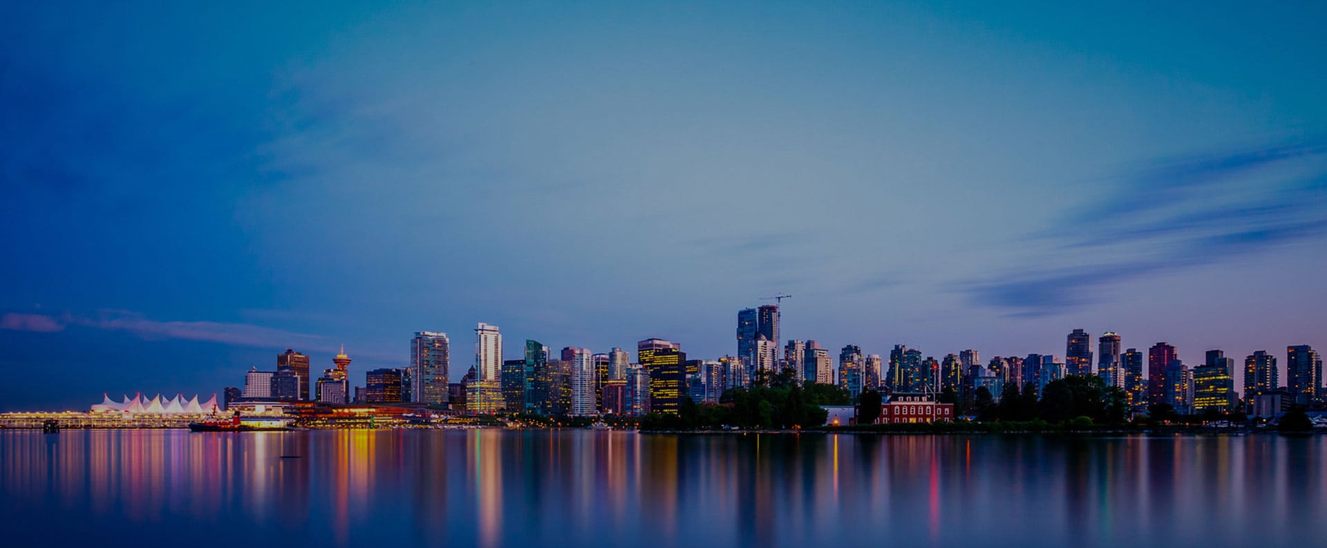 A large view of the Burrard Inlet waterfront at evening with lights from the skyline of Downtown Vancouver reflected on the waters.
